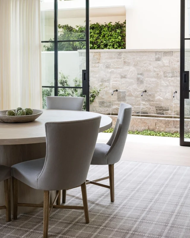 Framed by steel doors and soft linen drapery, the dining room at #mgdxlido feels equal parts refined and romantic. A custom pedestal table anchors the space, while light fabrics, a patterned wool rug, and the glow of the limestone fountain beyond create the dreamiest setting for gathering. #mindygayerdesign { Image by: @vlentine, Architect: @brandonarchitects, Builder: @longmanconstruction }